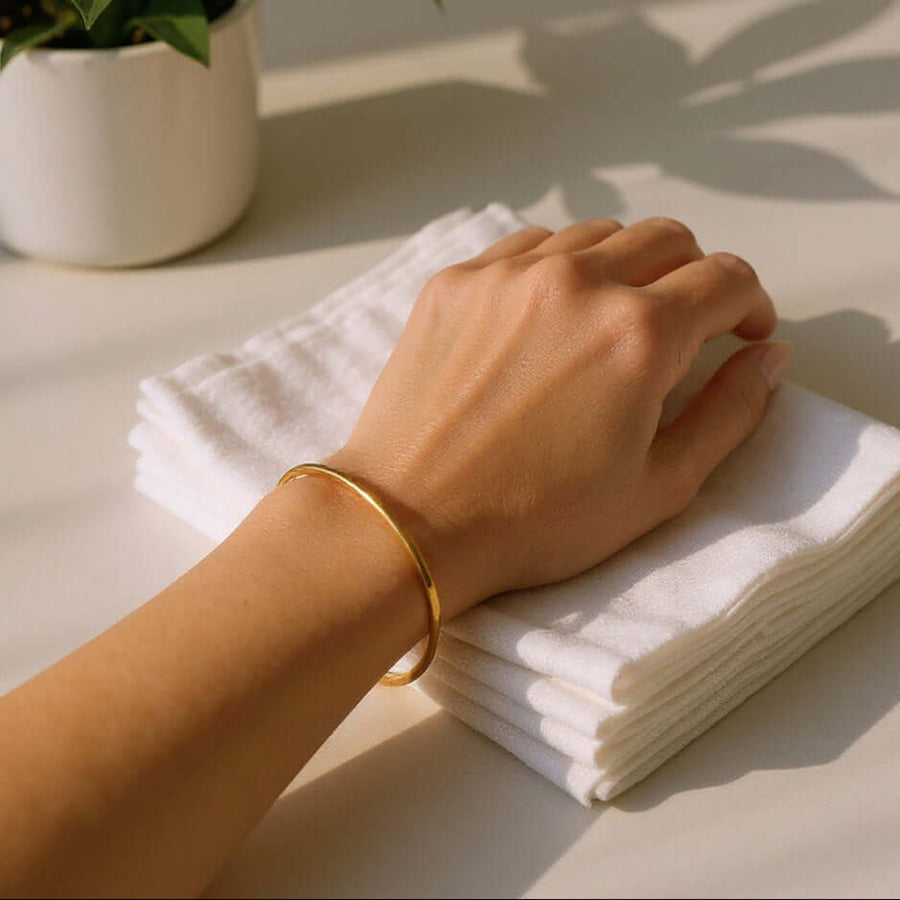 Hand with a gold bracelet on a stack of white napkins with a potted plant in the background.