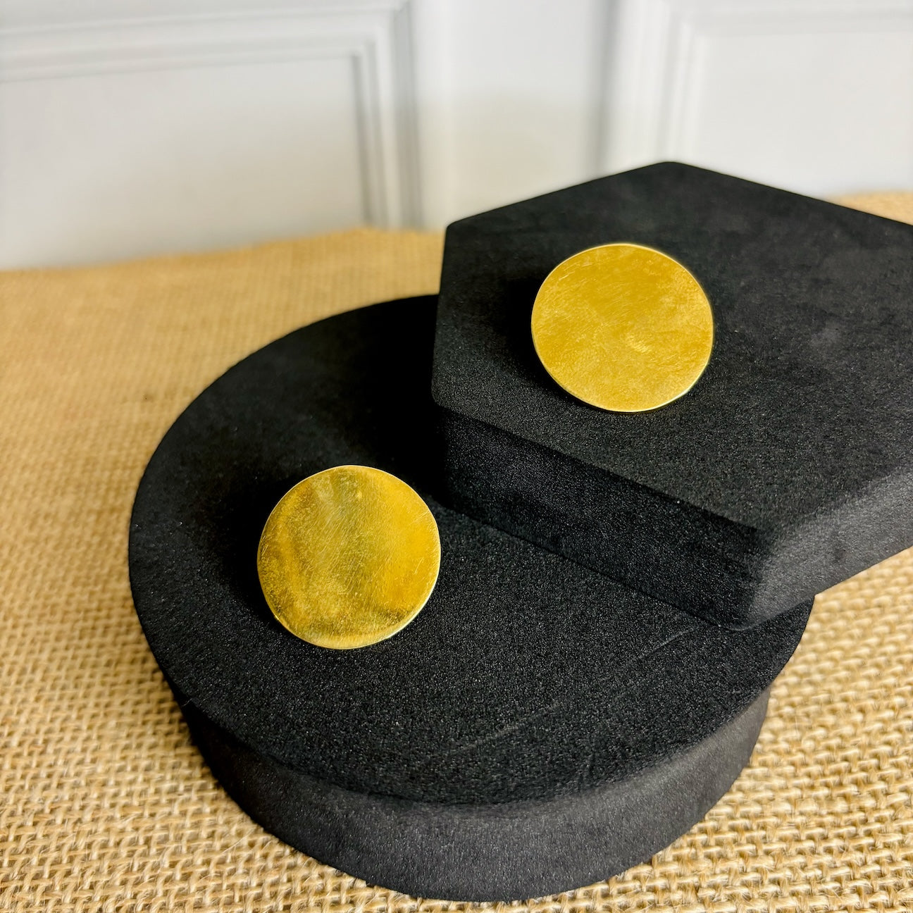 A pair of gold-colored round earrings displayed on a textured black stand, with the earrings' reflections visible on the stand.