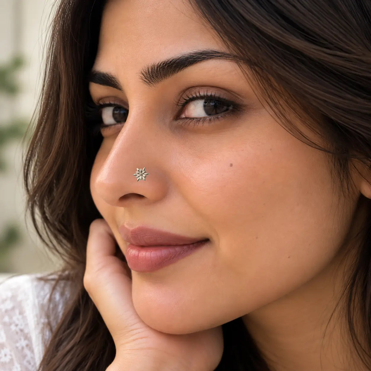 A close-up of a woman with a small yellow gold nose pin on her left nostril, resting her chin on her hand.