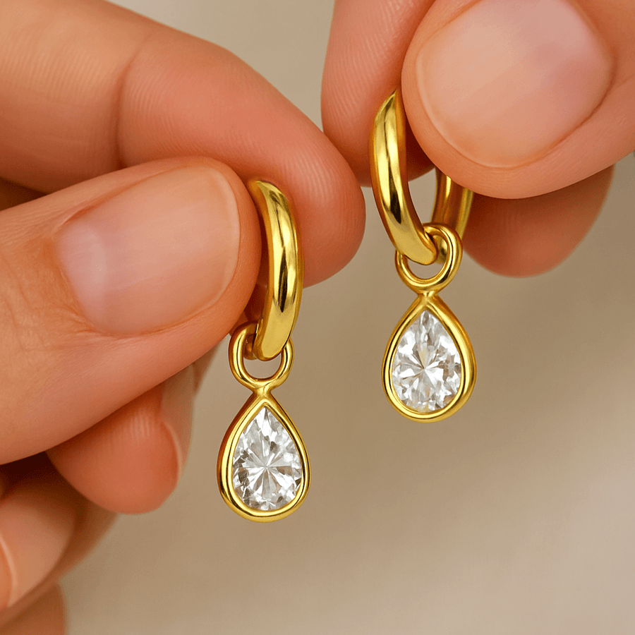 Gold earrings with teardrop-shaped gemstones held between fingers against a neutral background