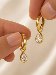 Gold earrings with teardrop-shaped gemstones held between fingers on a beige background