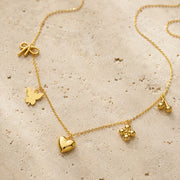 A delicate gold chain necklace with multiple small charms, including a bow, butterfly, heart, flower, and cherries, displayed on a textured beige stone surface.