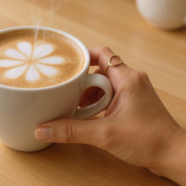 Hand holding a steaming cup of coffee with latte art on a wooden table wearing a gold ring