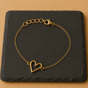 Mehr Heart Bracelet featuring a delicate open heart charm on a gold chain against a slate background.
