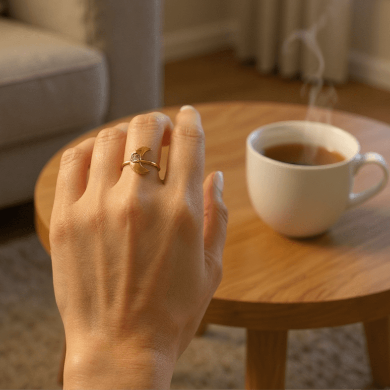 Hand with a Selena-Moon Charm Ring ring on a wooden table with a cup of coffee, blurred background